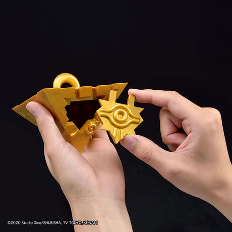 A pair of hands holding a gold-colored fantasy key-shaped accessory against a solid black background. The accessory has a triangular keyhole form with a ring at the top and a separate star-and-eye piece being inserted or aligned by the right hand.