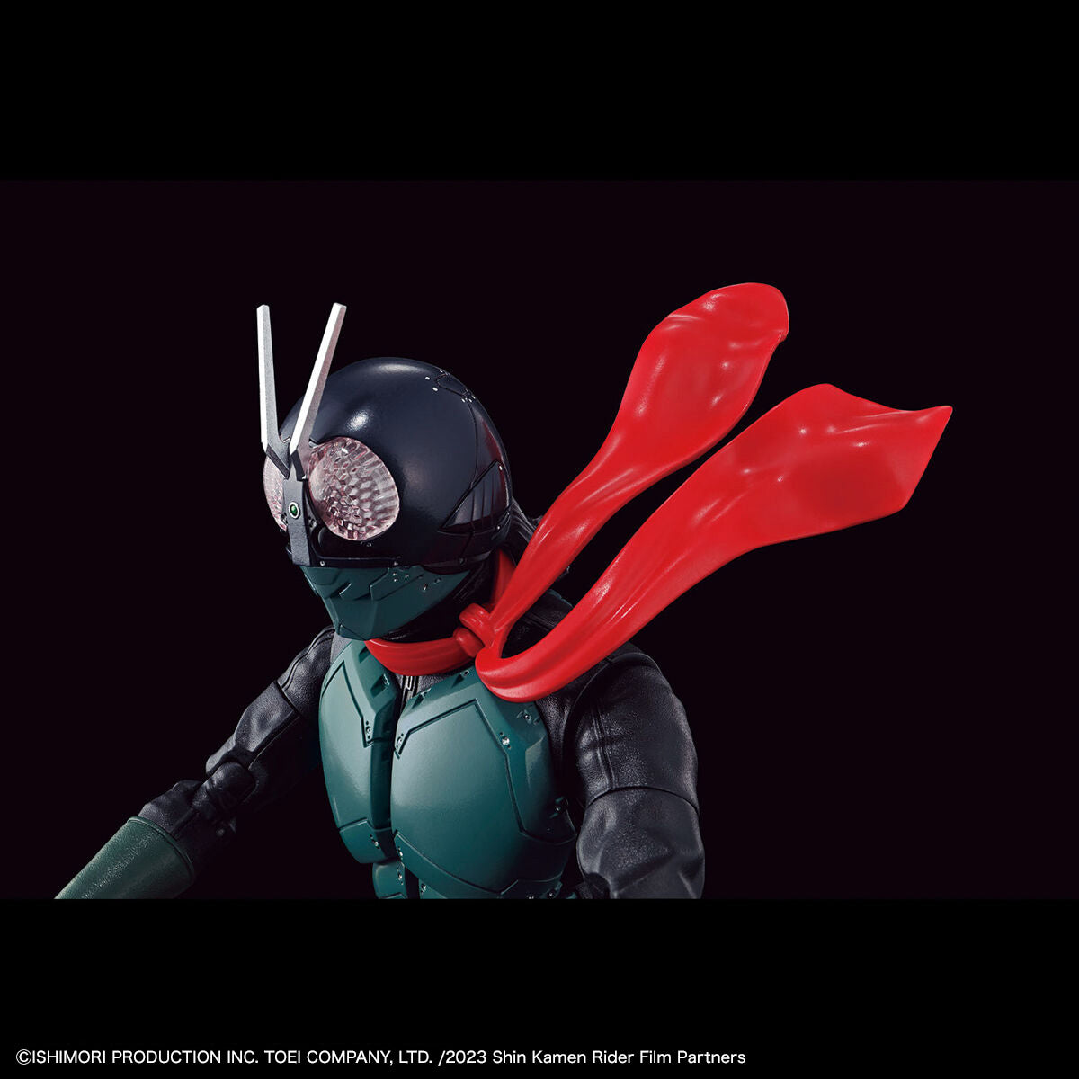 Shin Kamen Rider action figure or collectible character shown in a close-up studio shot against a black background, with a black helmet, teal chest armor, dark arms, and a bright red cape flowing behind it. Small copyright text appears along the bottom edge.