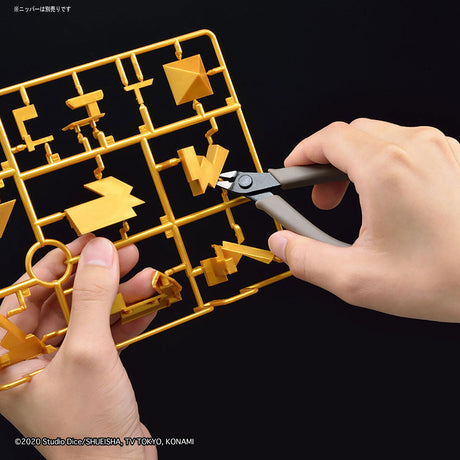 A close-up of a person's hands holding a yellow plastic model sprue and using metal wire cutters to trim a small yellow plastic piece, set against a solid black background.